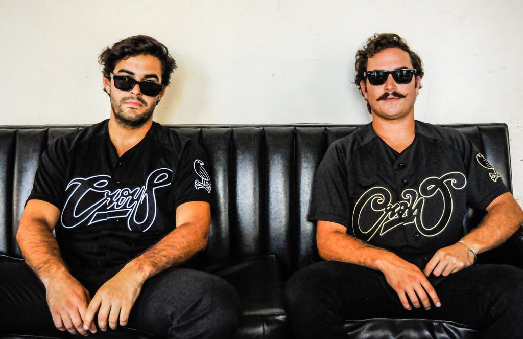 Two models wearing Crowdead White & Gold jerseys and sunglasses sitting leisurely on black couch. First image of collection.